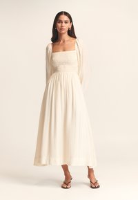 Cremefarbenes, langes Kleid mit gerafftem Oberteil und quadratischem Ausschnitt. Fließender Rock, lange Ärmel, aus leichtem Stoff gefertigt. Abgerundet mit schlichten Sandalen.