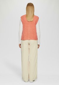 Rosa gestricktes, ärmelloses Oberteil, das über einem weißen Langarmshirt getragen wird, kombiniert mit lockeren, cremefarbenen weiten Hosen.