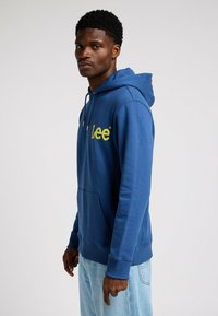 Sudadera de algodón azul con bolsillo tipo canguro y capucha con cordón. Presenta un logo amarillo de "Lee" en el pecho. Puños y dobladillo acanalados.