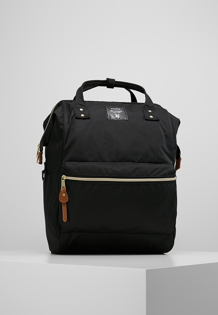 Sac à dos en toile noire avec deux poignées supérieures, poche avant zippée, accents en cuir et un patch logo distinctif à l'avant.