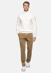 White knit turtleneck sweater, beige corduroy trousers, and white sneakers. Model stands with hands in pockets, facing forward.