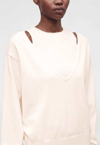 Personne portant un sweat-shirt crème avec des découpes uniques aux épaules et un design superposé en forme de V au niveau de l'encolure.