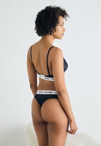 Zwarte kanten bralette met verstelbare bandjes, voorzien van een witte merkband. Bijpassende zwarte string met kanten rand. Glad textuur, minimalistisch ontwerp.