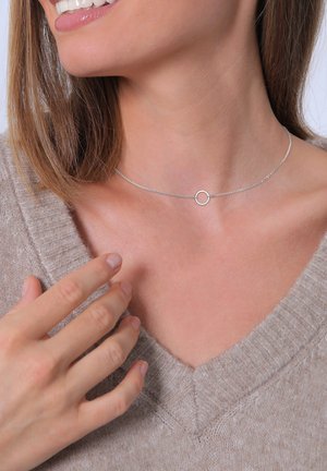 Silver choker necklace with a delicate chain and a circular accent. Worn close to the neck, paired with a light brown sweater.