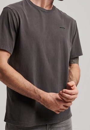 Homme portant un t-shirt à manches courtes gris foncé, les mains jointes devant, montrant un tatouage partiel sur le haut du bras gauche.