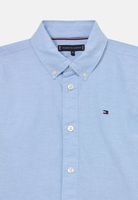 Camisa azul clara de botão com colarinho clássico, botões brancos e um pequeno logótipo bordado no lado esquerdo do peito. Fabricada em algodão.