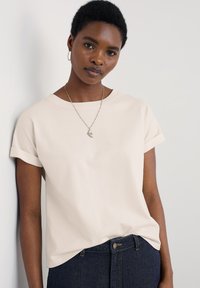 T-shirt beige a maniche corte con scollatura rotonda, maniche a risvolto e texture liscia, abbinato a jeans in denim scuri con chiusura a bottone.