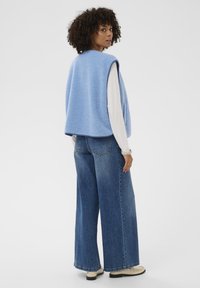 Frau mit lockigem Haar, die eine blaue ärmellose Fleeceweste, ein weißes Langarmtop, weit geschnittene blaue Jeans und beigefarbene Schuhe trägt, steht vor einem weißen Hintergrund.