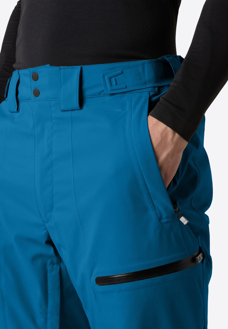 Ingrandimento di una persona che indossa pantaloni impermeabili blu con tasche laterali, cerniere e cintura regolabile, mano inserita nella tasca destra.