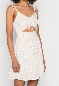 Šaty s květinovým potiskem, tenkými ramínky, uzlovým předním dílem a stupňovitými volány na spodním okraji. Mají bílý základ s oranžovožlutými květinami.