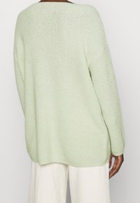 Personne portant un pull ample en tricot vert clair associé à un pantalon blanc, vue de dos sur un fond uni.