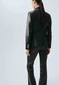 Černý kožený blazer a kalhoty, hladká textura, přiléhavý střih, se stříbrnými knoflíkovými detaily na rukávech blazera, pohled zezadu.