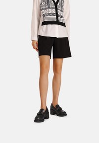 Mocassini neri con tacco blocco e decorazione a catena, indossati con pantaloni corti neri e una camicia bianca con motivi geometrici neri.