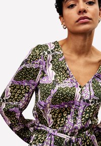 Frau in grünem Kleid mit lila Blumen- und Leopardendruck, V-Ausschnitt, Puffärmeln und tailliertem Bund, gezeigt von der Mitte des Oberkörpers bis zum Kinn.
