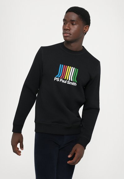 En ung mann iført en svart hettegenser med fargerike vertikale striper og "PS Paul Smith"-logo, som ser mot venstre mot en hvit bakgrunn.