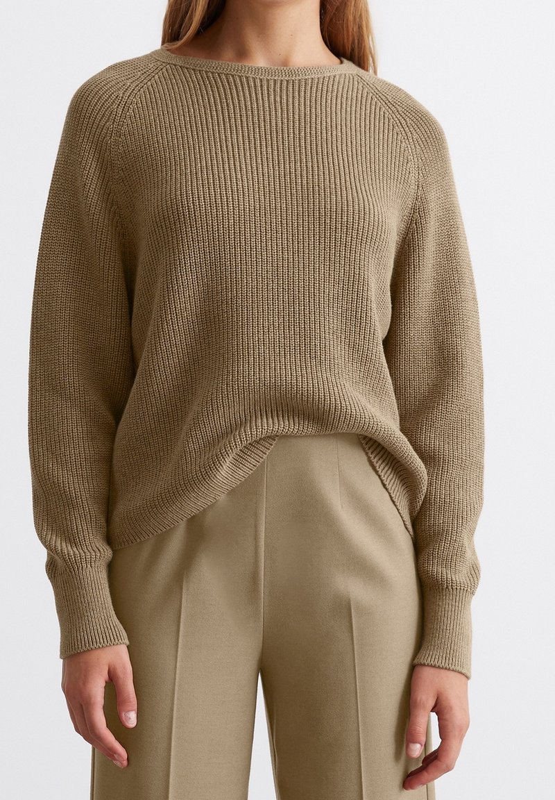 Femme portant un pull en maille côtelée beige légèrement rentré dans un pantalon beige taille haute et ajusté, les mains détendues le long du corps.