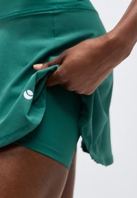 Roka paceļ zaļu sporta svārku, atklājot tam atbilstošus zaļus šortus ar nelielu baltu logotipu svārku apakšmalā.