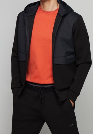Uomo con giacca nera con zip e cappuccio sopra una maglietta arancione girocollo e pantaloni neri con coulisse, mani nelle tasche.