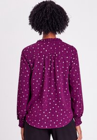 Blouse mauve à manches longues avec un col à volants, ornée de motifs en diamants argentés et de poignets élastiqués. Tissu lisse avec une coupe décontractée.
