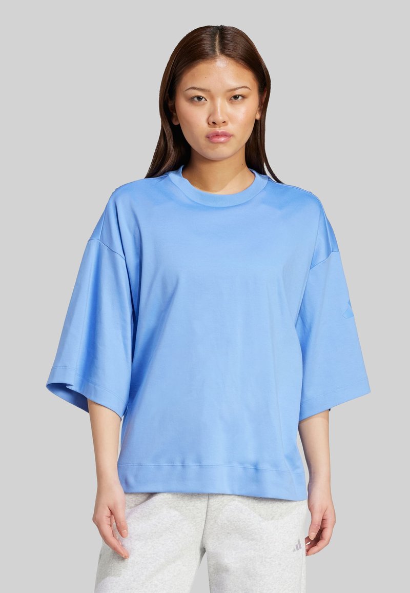 Modrá oversized tričko z jemnej bavlny, s okrúhlym výstrihom, spadnutými ramenami a krátkymi rukávmi. Hladká textúra, minimálny dizajn.