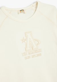 Krämfärgad bomullströja med broderad text "A" och "THE ACADEMIC CLUB EST.1996" i beige. Har rund halsringning och raglanärmar.