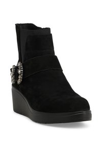 Stivaletto alla caviglia in suede nero con tacco a zeppa, caratterizzato da una cinturina decorativa e un accento di strass sul lato. Tessitura liscia e design minimalista.