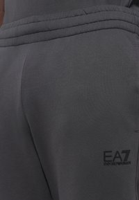 Šedé tepláky vyrobené z jemnej tkaniny, s elastickým pásom na šnúrku a malým logom EA7 na boku. Hladký povrch bez vzorov.