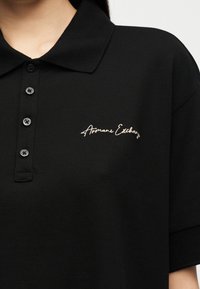 Μαύρο πόλο μπλουζάκι με κοντά μανίκια, γιακά, τρία μαύρα κουμπιά και λευκό κεντητό λογότυπο "Armani Exchange" στην αριστερή πλευρά του στήθους.