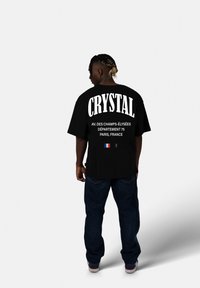 Černé oversized tričko s velkým bílým nápisem "CRYSTAL" a detaily o lokaci pod ním. V kombinaci s tmavými džínami a teniskami.