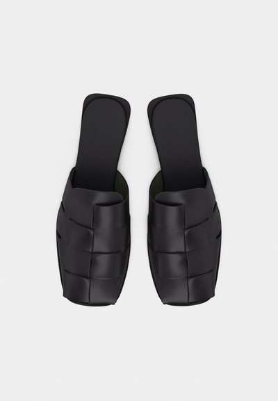 Μαύρα σανδάλια τύπου slip-on με πλεκτό δερμάτινο σχέδιο και επίπεδες τετράγωνες σόλες, εμφανιζόμενα από πάνω σε λευκό φόντο.