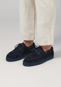 Navy blue suede shoes with a rounded toe, lace-up design, and woven rope sole; paired with cream-colored pants.