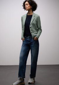 Blazer vert clair texturé avec boutons noirs, haut bleu marine, jean bleu décontracté et baskets grises. Le mannequin pose les mains dans les poches.