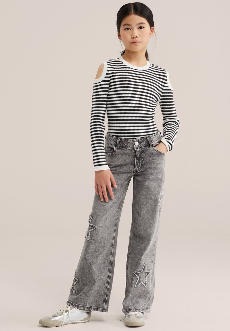 Jeune fille debout, la main dans la poche, portant un top rayé noir et blanc à épaules dénudées, un jean gris à larges jambes avec des étoiles en patch, et des baskets blanches.