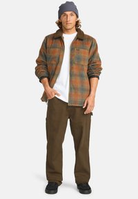 Geruite flanellen shirt in oranje, groen en blauw met een bruine kraag; gecombineerd met bruine broek en zwarte schoenen. Model draagt een grijze beanie.