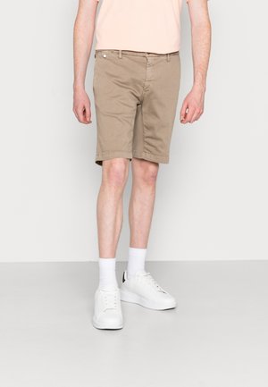 Mann trägt beige knielange Shorts, weiße Socken und weiße Sneakers, steht vor einem einfarbigen hellen Hintergrund.