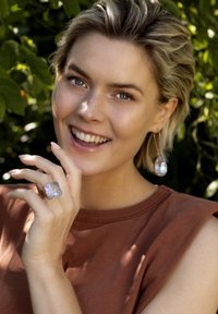 Gouden ring met een grote, transparante, iriserende steen. Bijpassende druppeloorbellen hebben soortgelijke stenen. Model met kort, tousled haar en een bruine top.