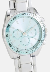 Montre-bracelet en métal argenté avec un cadran bleu clair, trois sous-cadrans et des marquages noirs. Elle est dotée d'un bracelet lisse et poli ainsi que d'index des heures audacieux.