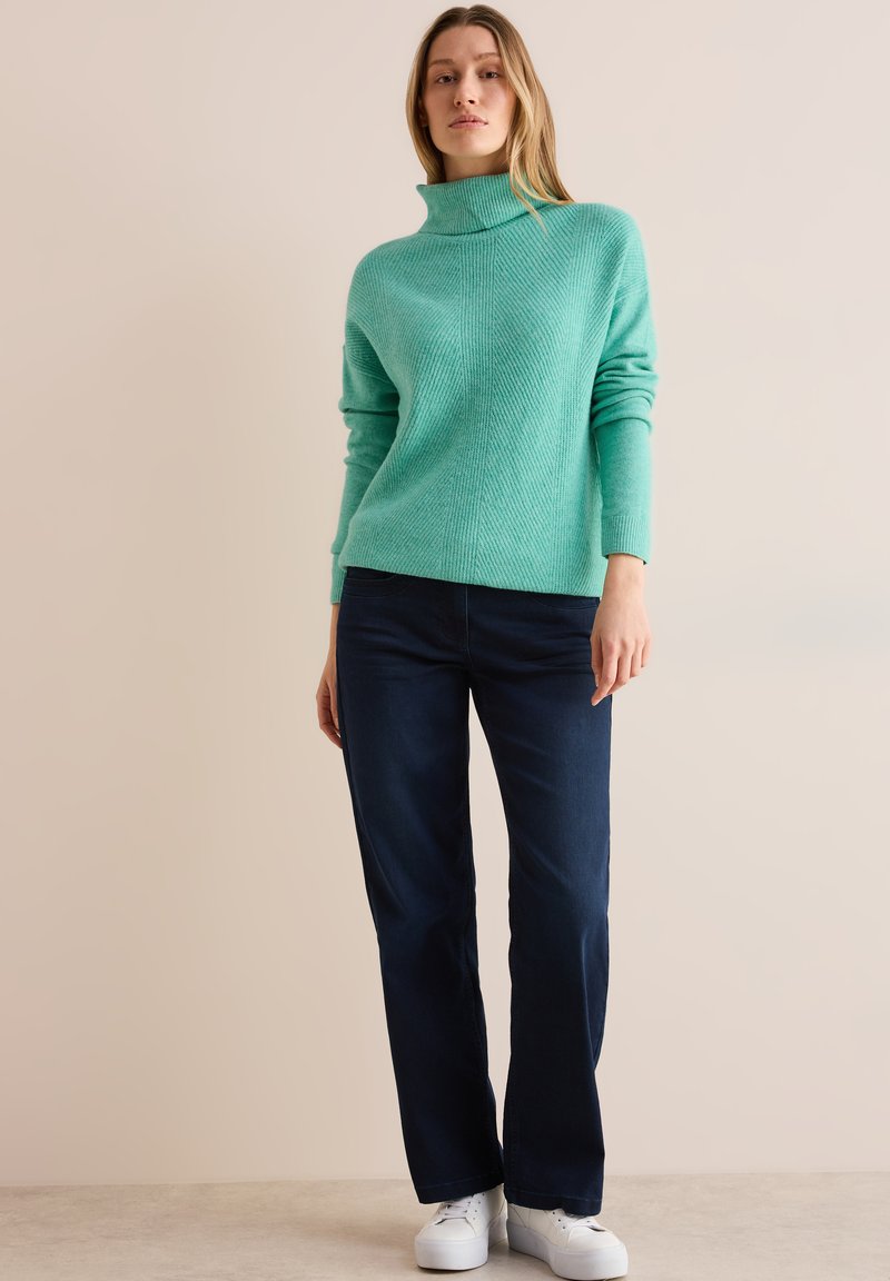 Suéter de cuello alto turquesa con un patrón texturizado, combinado con jeans rectos azul oscuro y zapatillas blancas. Atuendo simple y casual.