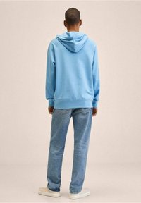 Uomo in piedi di spalle, vestito con un felpa leggera blu, jeans blu e scarpe bianche, su uno sfondo beige uniforme.