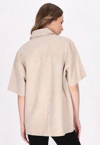 Camicia beige in pelle a maniche corte con taglio dritto, dettagli di cucitura visibili e colletto classico, vista dal retro.