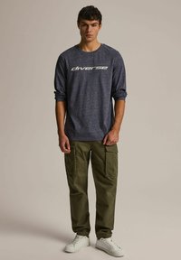 Granatowa koszula z długim rękawem z logo "diverse", w połączeniu z oliwkowymi spodniami cargo z wieloma kieszeniami, noszona z białymi trampkami.