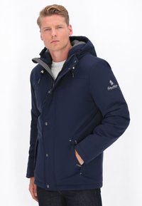 Marineblau isolierte Jacke mit Reißverschluss und Druckknopfverschluss, graue Fleece-gepolsterte Kapuze, seitliche Taschen und einem Logo auf dem Ärmel.