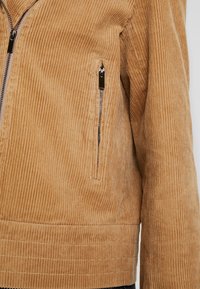 Kurtka z korduroy w ciepłym odcieniu brązu, z pionowymi żeberkami, kieszeniami zapinanymi na zamek oraz dopasowanym talią z poziomymi detalami szwów.