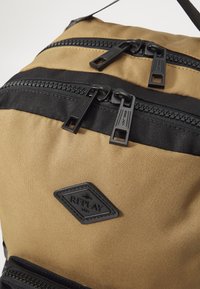 Tan nylon backpack featuring two black zippers, black accents, and a diamond-shaped logo patch with the text "REPLAY 1991." Textured surface.