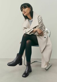 Ghete negre din piele până la gleznă, cu vârf pătrat și toc bloc. Prezintă părți elastice și o textură netedă. Purtați cu colanți negri și un trench.
