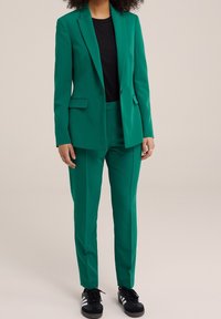 Žena v zeleném sešitě na míru s černou košilí a černými teniskami Adidas se bílými pruhy, stojící proti neutrálnímu pozadí.