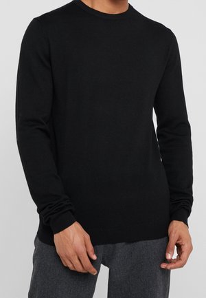 Mann in einem schlichten schwarzen Langarmpullover und dunkelgrauen Hosen, der mit entspannten Händen vor sich steht.