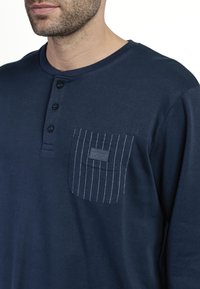 Camisa de manga larga azul marino con un cuello abotonado, que presenta un bolsillo en el pecho izquierdo con rayas verticales blancas y una etiqueta con nombre.