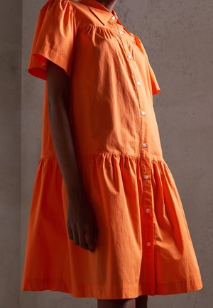Personne portant une robe orange ample à manches courtes et boutonnée, avec une jupe froncée, debout devant un fond uni.
