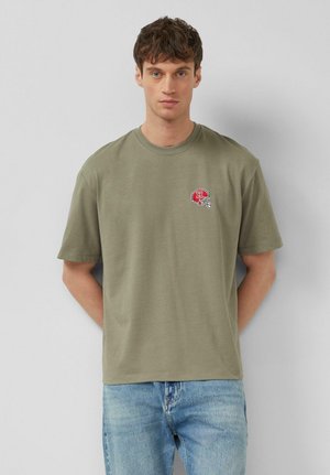 Ragazzo giovane con capelli corti e scuri che indossa una t-shirt verde oliva con un piccolo logo di un casco rosso e jeans azzurri chiari, in piedi davanti a un muro grigio.